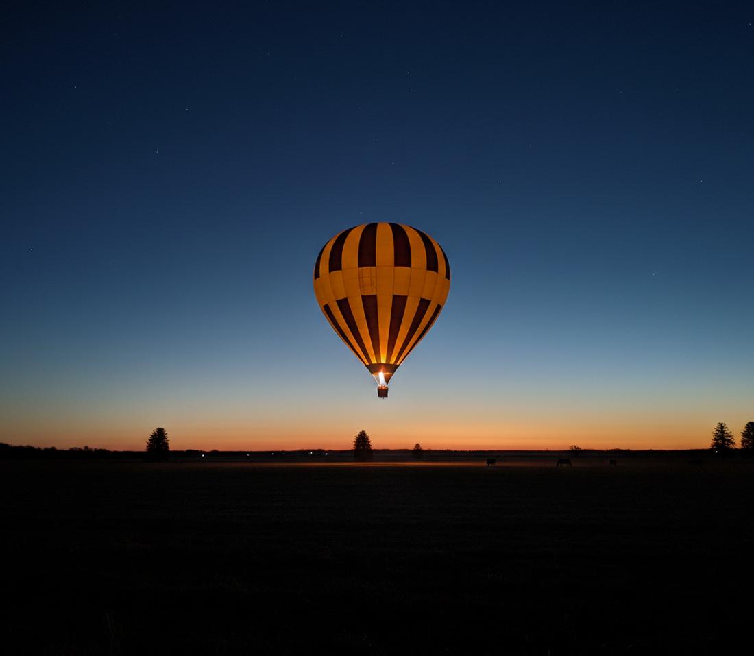 A striped yellow-orange and brown balloon lit from inside by its burner in the navy sky before dawn, a thin band of first light on the horizon and horses in silhouette